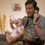 Telefonerende vader met huishoudschort geeft baby de fles. Nederland, [1961]