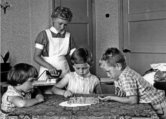 Terwijl de kinderen een potje ‘Mens Erger Je Niet’ spelen, staat een jonge gezinshulp te strijken, 1958. (Nationaal Archief/Spaarnestad/Pim Stuifbergen)
