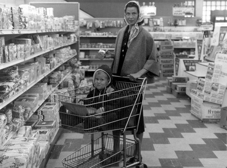 Huisvrouw met hoofddoek om, met haar zoontje in winkelwagen, doet boodschappen in de supermarkt. (Nationaal Archief - wiki)