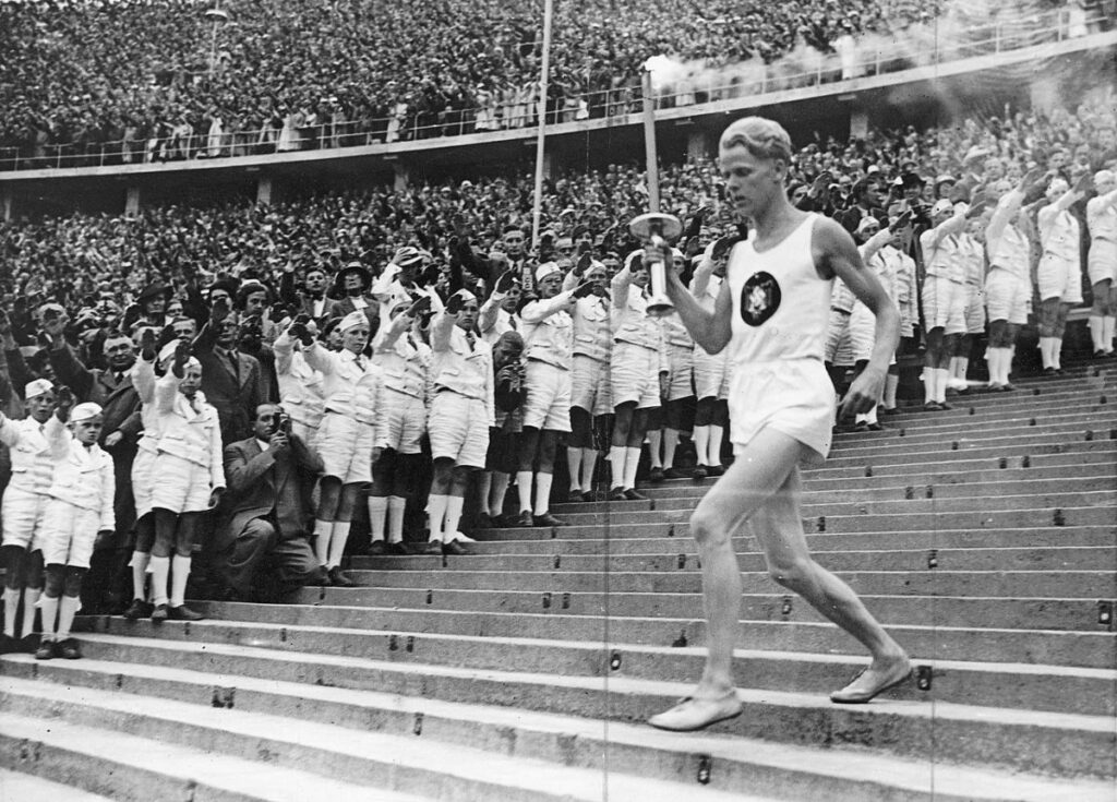 Fritz Schilgen loopt het stadion binnen met de fakkel, onder toeziend oog van leden van de Hitlerjugend