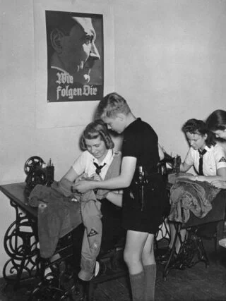 BDM-meisjes naaien kleding. Aan de wand een portret van Adolf Hitler