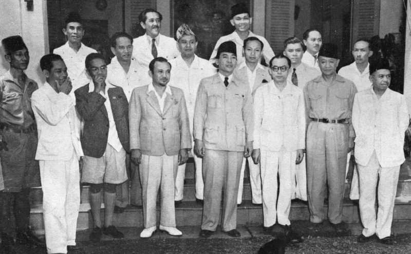Het eerste kabinet van de Republiek Indonesië. In het midden Soekarno, rechts naast hem met bril Hatta. Op de voorste rij tweede van links (korte broek en kousen) Amir Sjarifoeddin. 