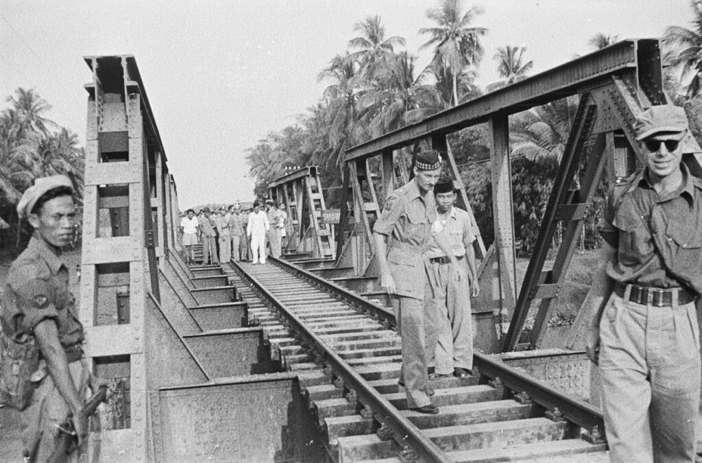 Een Britse militaire waarnemer op 26 april 1948 aan de status quo-lijn bij Gombong (Midden-Java). Op de achtergrond Indonesische militairen, op de voorgrond Nederlandse. Midden op de brug aan de rechterkant een bord met de tekst Status Quo. 