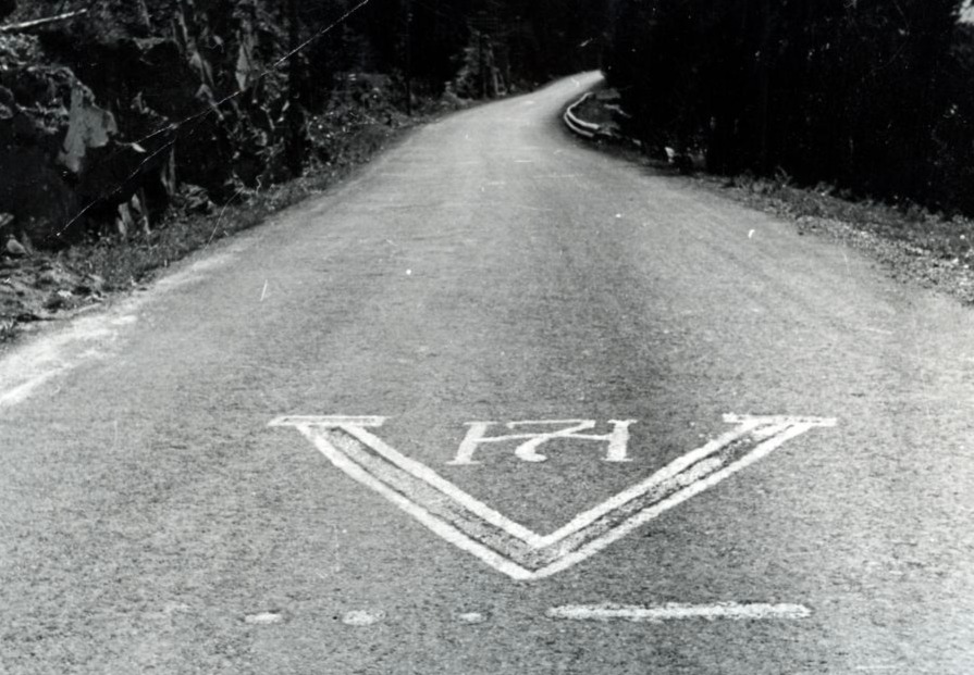 Het V-teken op een weg in Noorwegen, aangebracht door leden van het verzet. Onderaan de 'V' in morsecode en bovenaan het monogram van de Noorse koning Haakon VII.