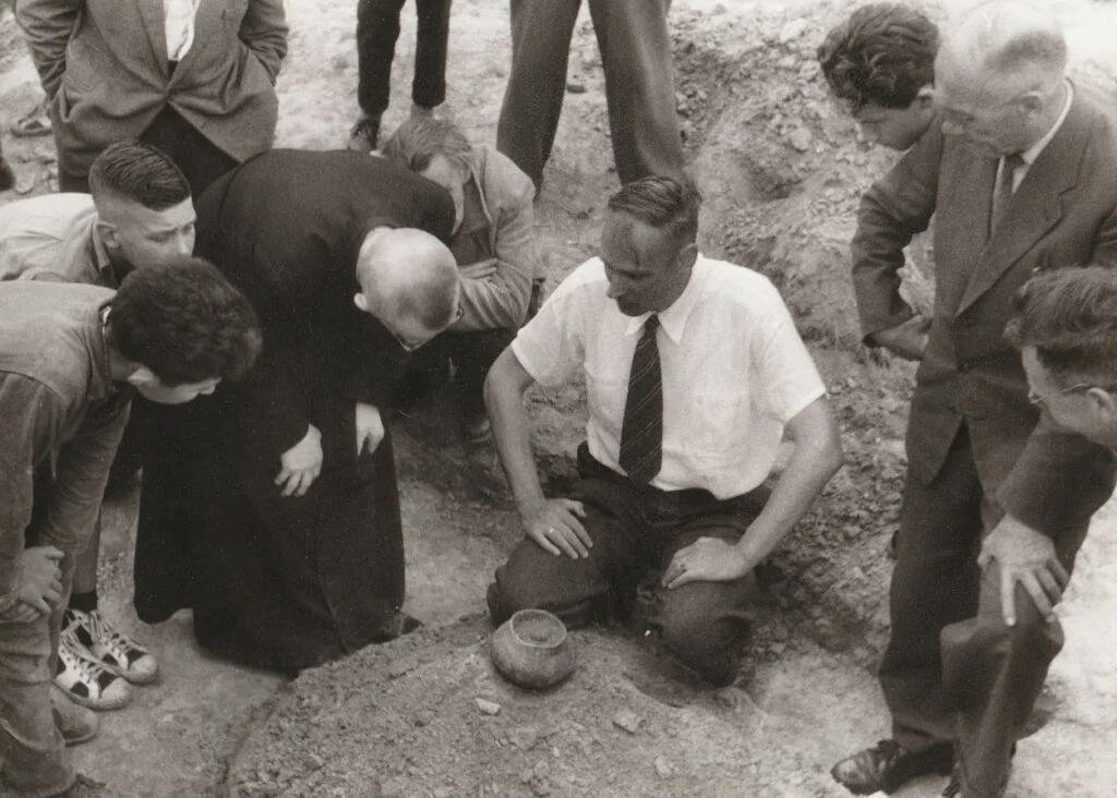 Prof. dr. Modderman, zittend te midden van vele toehoorders geeft aan de lokale geestelijke uitleg over de bandkeramische graven van Elsloo, 1966
