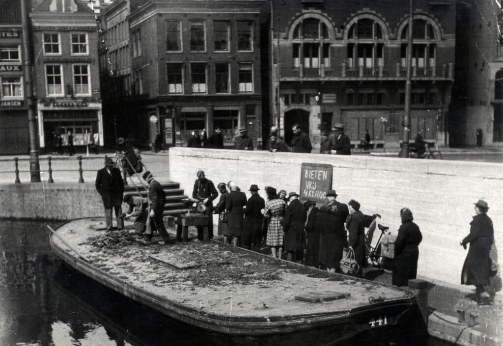 Hongerwinter WO II. Op en naast een schuit op het water van het Rokin in Amsterdam wordt groente verkocht, waaronder bieten. Een lange rij inwoners van Amsterdam staat op de kade te wachten om wat voedsel te bemachtigen. Nederland, Amsterdam, 1945.