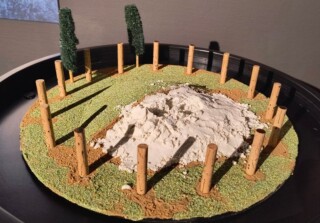 Miniatuur grafheuvel met de kenmerkende krans van palen aan de voet